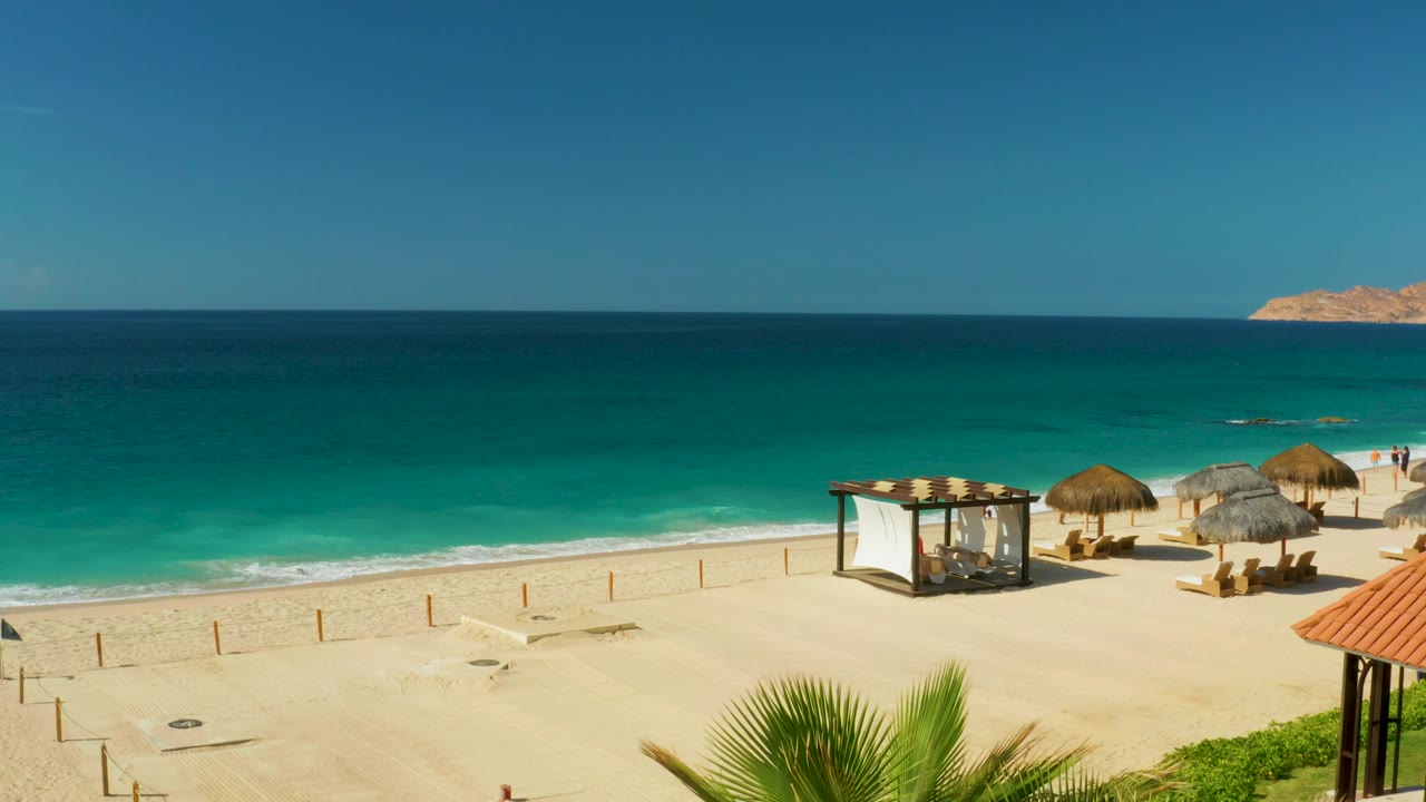 Hilton Los Cabos beach frame showing the ocean horizon, pale sand, a white canopy structure, several thatched umbrellas, lounge chairs, and no visible people.