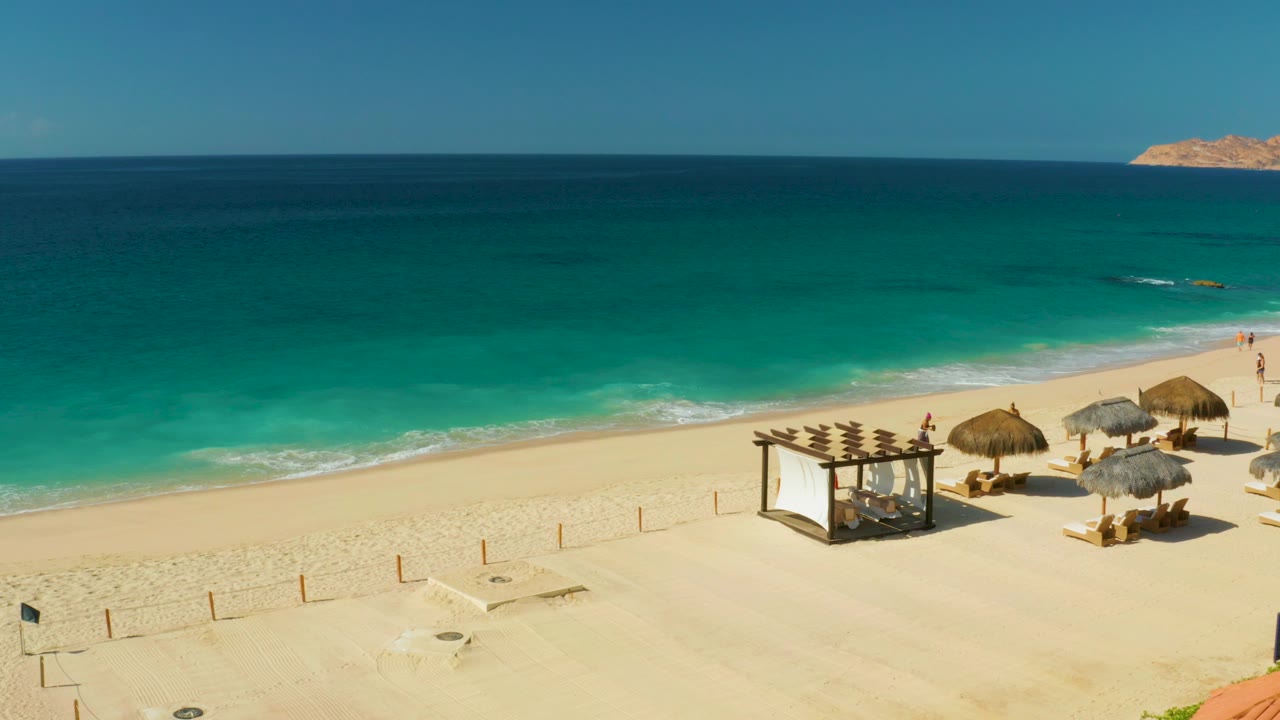 High aerial view at Hilton Los Cabos showing turquoise ocean, pale sand, thatched umbrellas, lounge chairs, a shade frame, and small beachgoers near the shoreline.