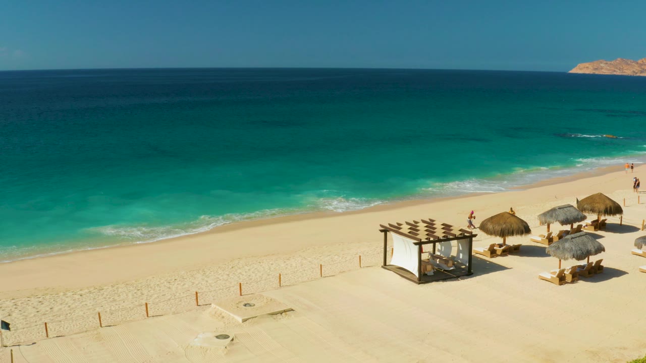Elevated Hilton Los Cabos view with deep blue water beyond turquoise shallows, pale sand, thatched umbrellas, lounge seating, and a few small people along the shore.
