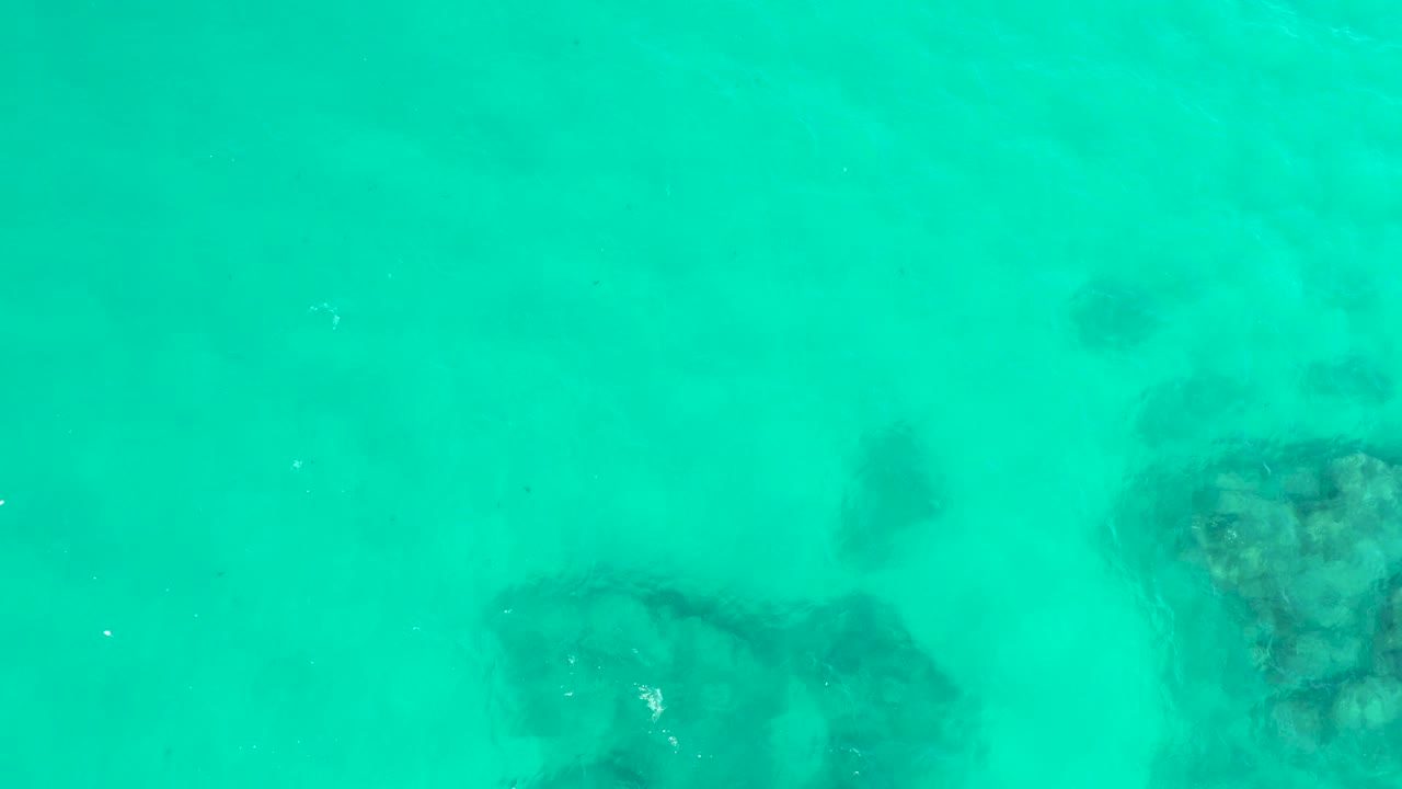 At Hilton Los Cabos in Mexico, turquoise water reveals a coral-like rock patch to the right and a softer dark patch to the left, with no visible people.