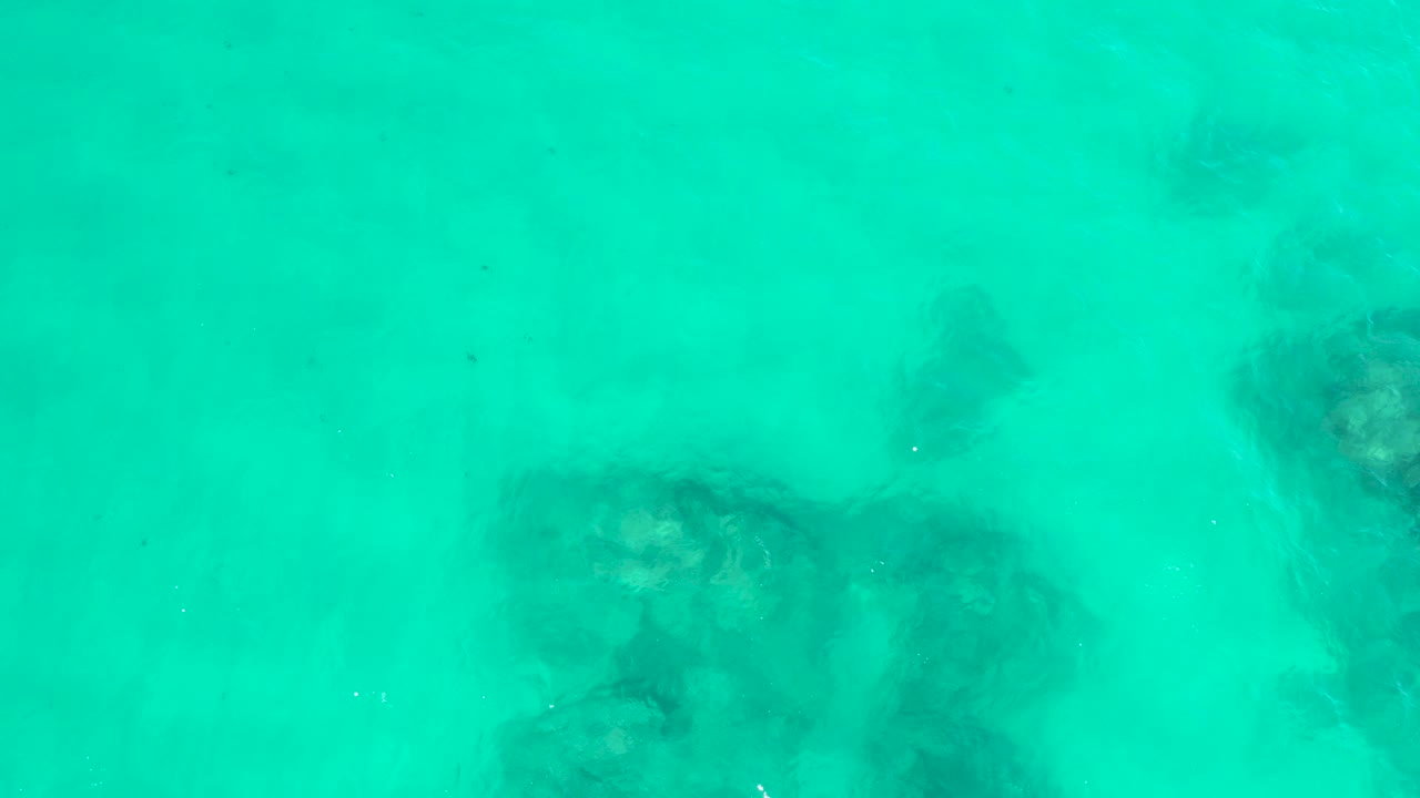 Overhead view of clear turquoise water at Hilton Los Cabos, Mexico, with dark submerged rock patches visible near the lower left and no visible people.