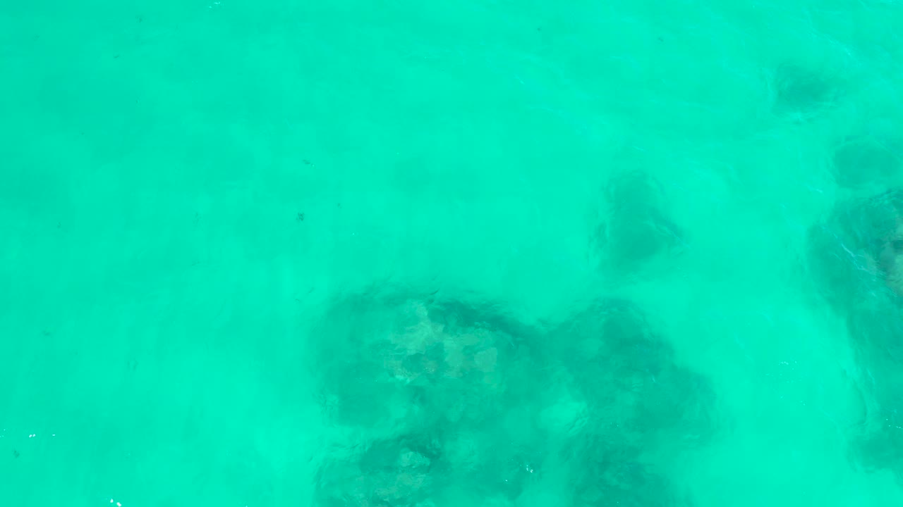 A top-down frame from Hilton Los Cabos, Mexico, shows turquoise water with submerged rocks across the lower half and a smaller dark patch near the upper center, with no visible people.