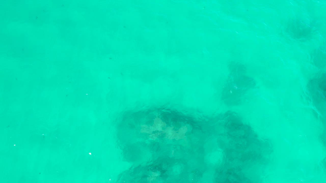 At Hilton Los Cabos, Mexico, the overhead view holds on pale turquoise water where a large darker submerged patch sits lower left and faint rock detail appears through ripples, with no visible people.