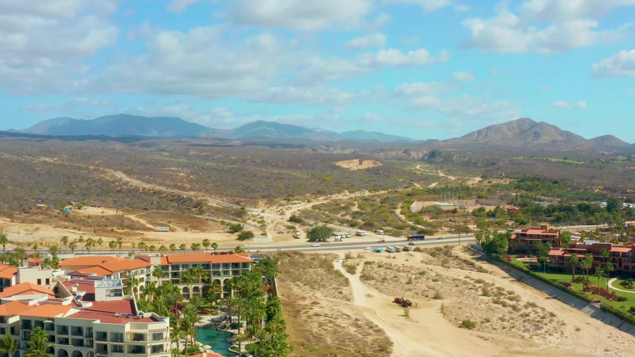 Aerial frame near Hilton Los Cabos shows the highway cutting across the midground, several small vehicles, red-roofed buildings in the foreground, low hills, and no visible people.
