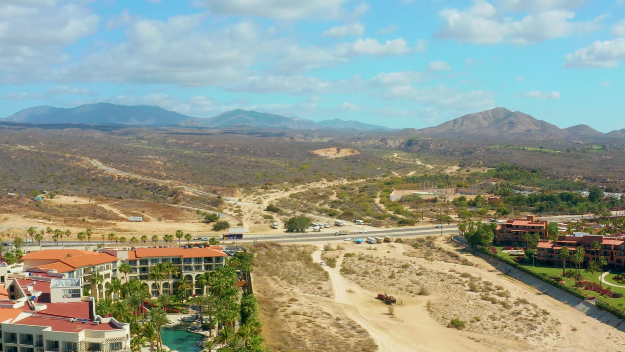 Aerial opening frame over Hilton Los Cabos, Mexico with red-roofed buildings and a pool at lower left, a road with cars across the midground, dry hills, distant mountains, scattered clouds, and no visible people.
