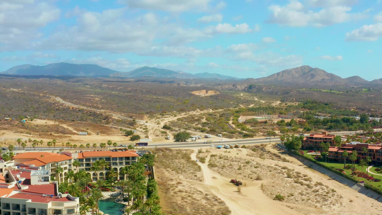 Aerial frame over Hilton Los Cabos, Mexico showing cars along the road, red-roofed resort buildings at the lower edge, scrub-covered desert hills, distant mountains, partly cloudy sky, and no visible people.