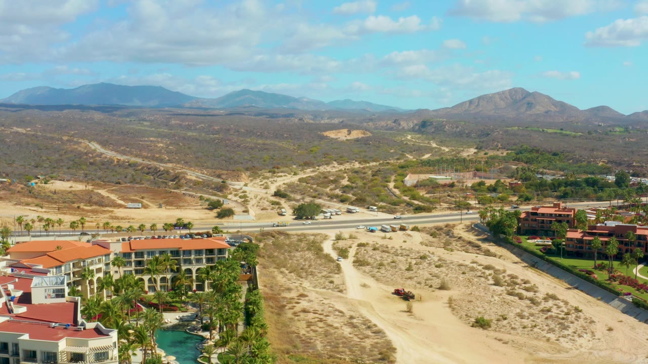 Aerial frame over Hilton Los Cabos, Mexico showing more of the resort rooftops and pool at the bottom, a line of cars on the road, pale dirt paths through dry terrain, mountain ridges, scattered clouds, and no visible people.