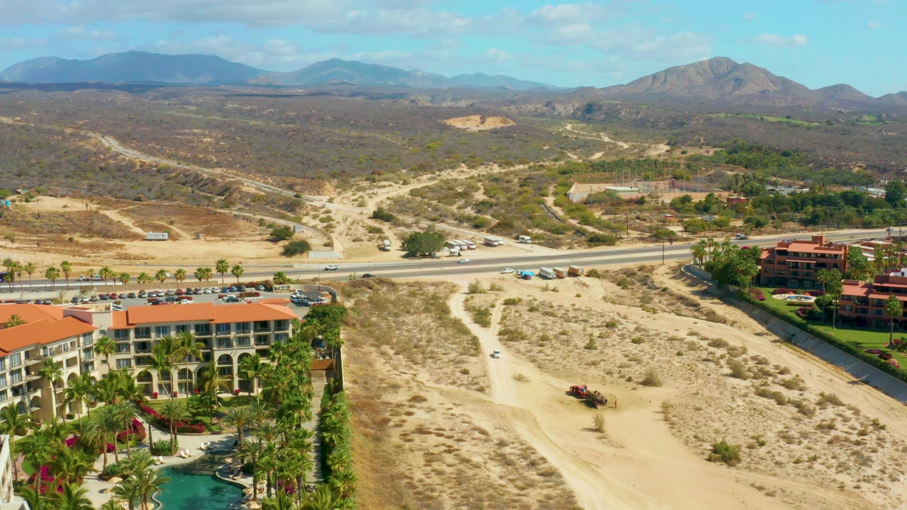 Aerial frame at Hilton Los Cabos, Mexico, with the orange-roofed resort and palms lower in frame, parked cars along the road, branching dirt paths through scrubland, mountains in the background, and no visible people.