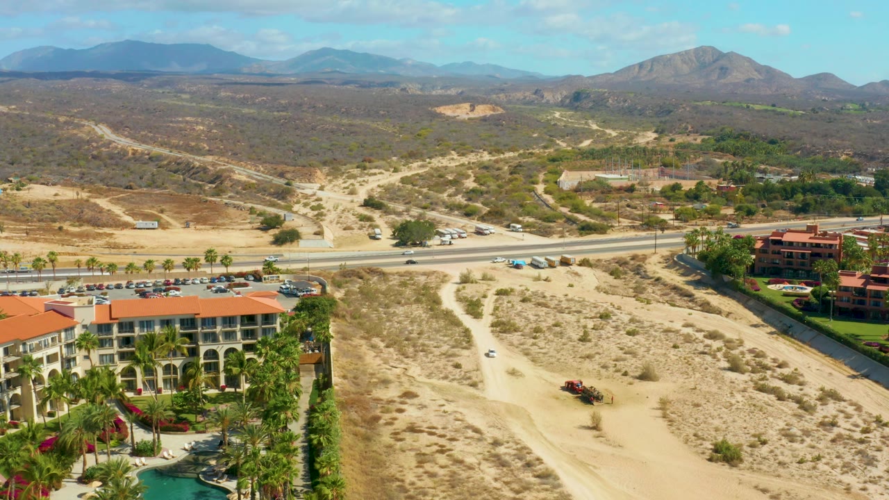 Mid-clip aerial view at Hilton Los Cabos, Mexico, showing the resort complex with palm landscaping, a road with small cars, sandy open terrain, winding dirt tracks, distant mountains, and no visible people.