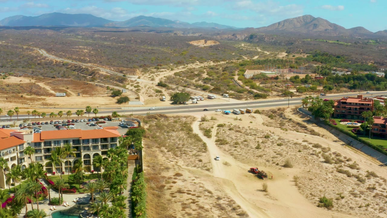 Wide aerial landscape with resort corner, road, dirt paths, parked cars, and mountains.