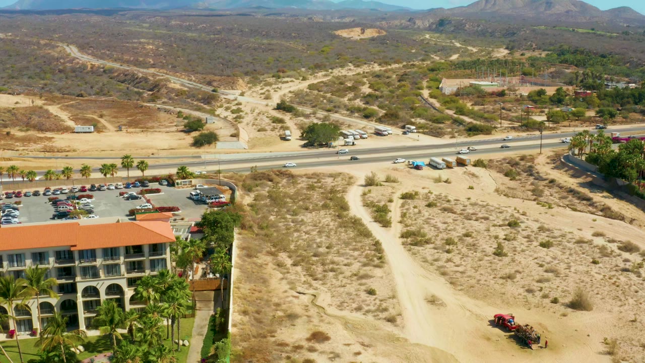 Second aerial frame at Hilton Los Cabos, Mexico with the hotel and palms lower left, pale dirt road crossing the desert, vehicles on the highway, mountains on the horizon, and no visible people.