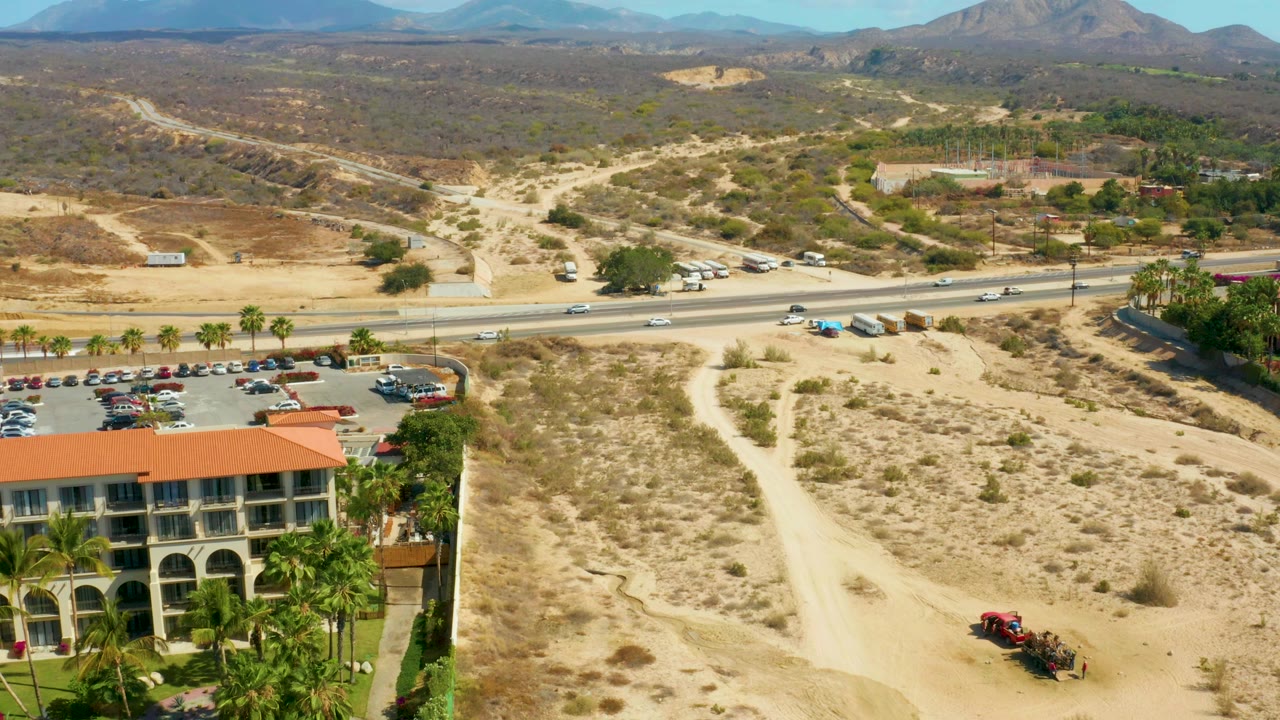 Third aerial frame at Hilton Los Cabos, Mexico showing a broader right-side desert slope, the hotel roof along the bottom, highway traffic in the midground, distant mountains, and no visible people.