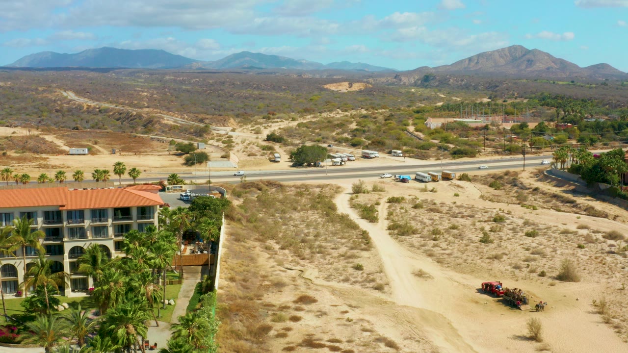 At Hilton Los Cabos in Mexico, an elevated view shows a red-roofed building with palms at left, a pale dirt path through dry scrub, road traffic beyond it, distant mountains, and no visible people.