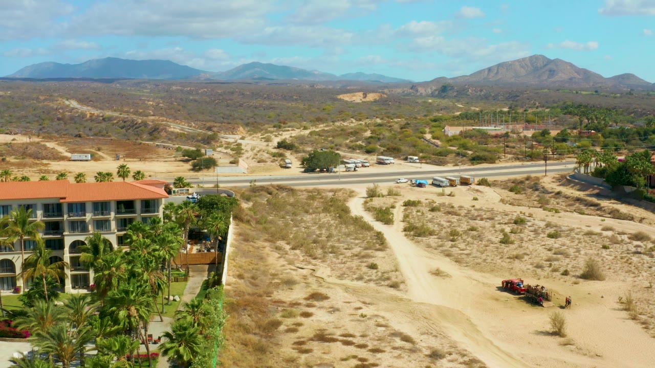 At Hilton Los Cabos in Mexico, the red vehicle sits farther along the lower dirt path while the red-roofed building, palms, paved road, desert scrub, and mountain horizon remain visible, with no visible people.