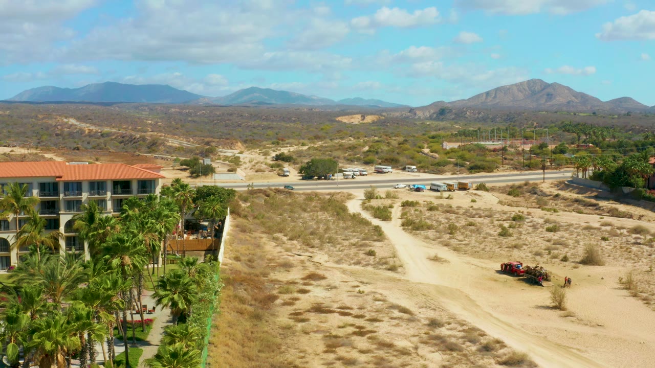 Desert roadway with building, palms, moving vehicles, and mountains under scattered clouds.