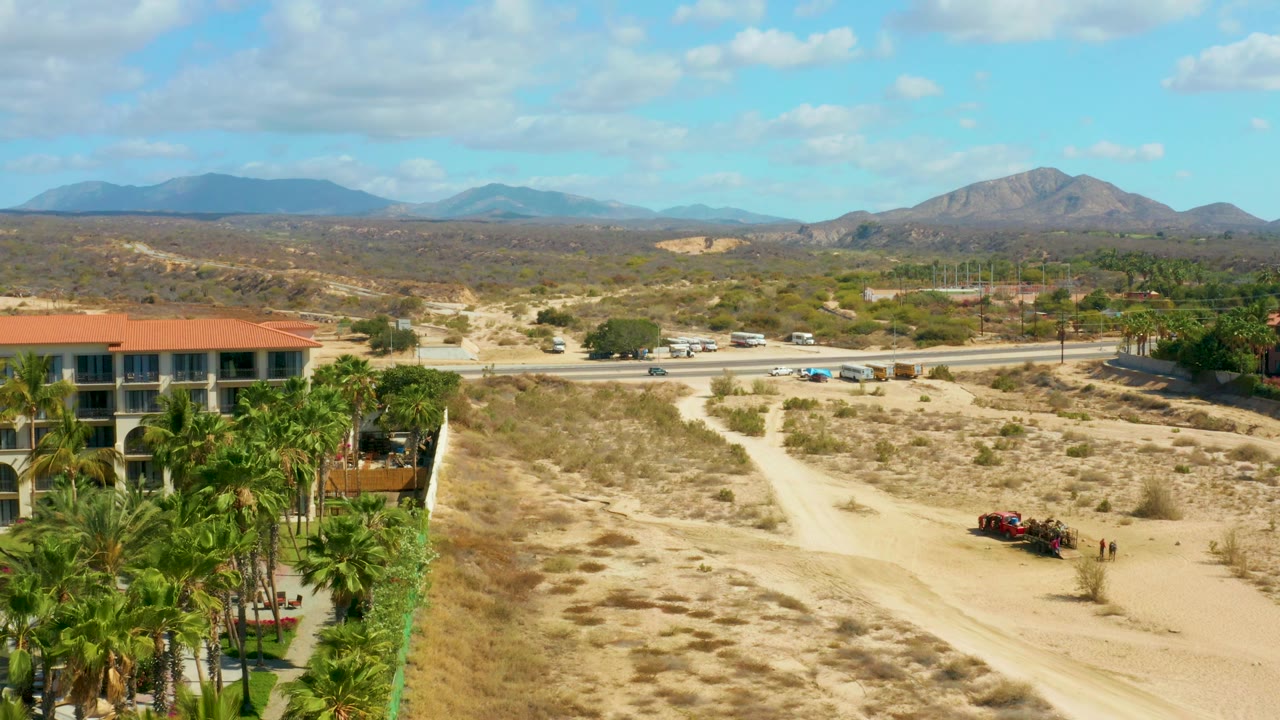 Aerial Hilton Los Cabos view with the sandy track running through the foreground, palm landscaping and a building at left, roadside vehicles across the middle, distant mountains, and no visible people.