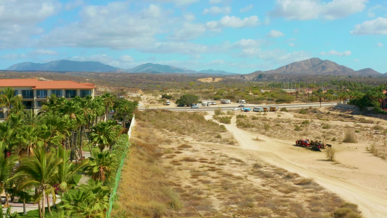 Opening frame at Hilton Los Cabos BCS Mexico showing a pale dirt path through dry grass, palms along the left edge, a red-roofed building, distant mountains, and no visible people.