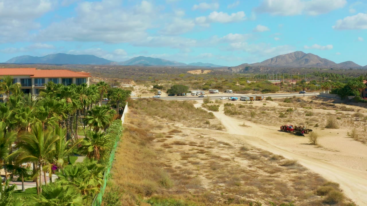 The cart remains on the right side of the dirt track while palm fronds fill more of the foreground edge, with the red-roofed building, parked vehicles, mountains, and no visible people.