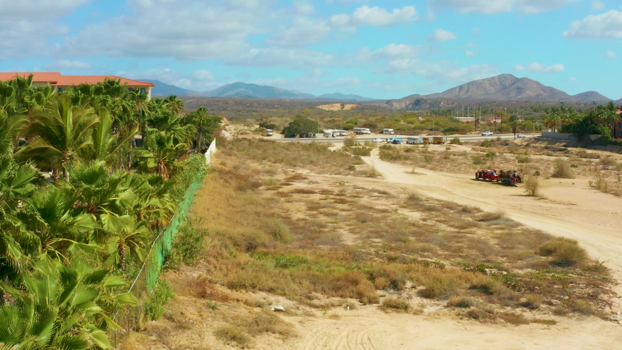 At Hilton Los Cabos, Mexico, a sandy dirt road runs past a green fence and palm trees toward distant mountains, with a small red vehicle on the road and no visible people.