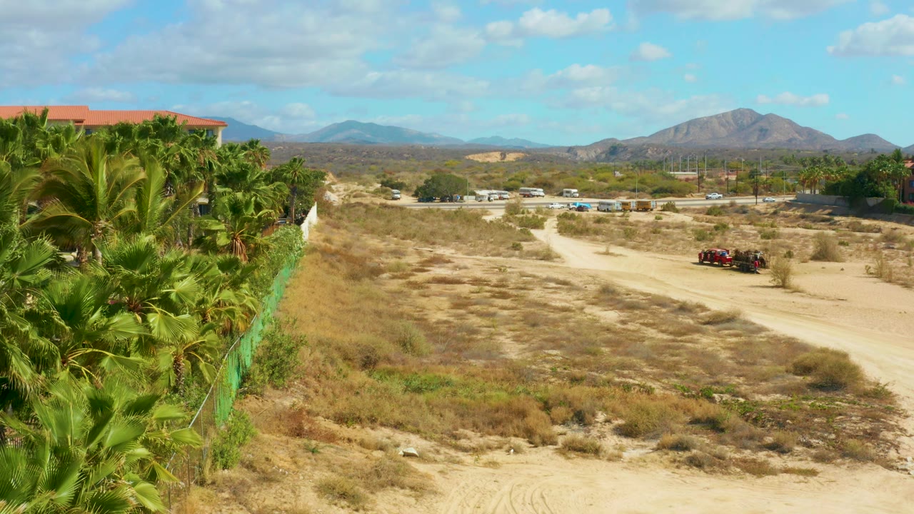 The elevated view holds the palm row and green fence at left while the pale dirt road and red vehicle sit farther right against distant hills, with no visible people.