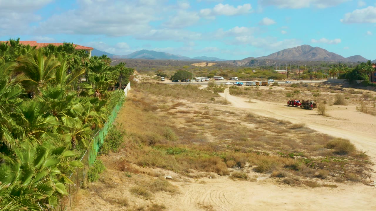 A green fence and thick palms fill the left side as the dry open area and dirt road extend toward a red vehicle and mountain horizon, with no visible people.
