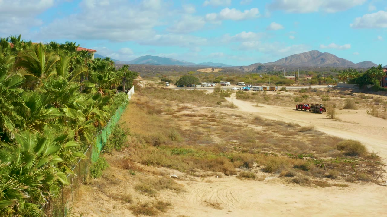 At Hilton Los Cabos, Mexico, the camera position shows more of the palm-lined fence on the left and the red vehicle near the right side of the road, with mountains and no visible people.