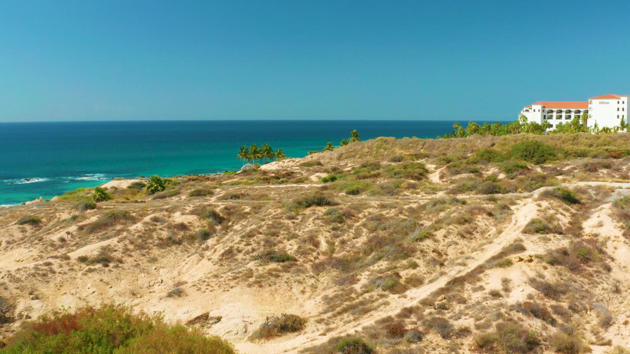 At Hilton Los Cabos BCS Mexico, dry scrub hillside and a pale dirt path sit below a blue ocean horizon, with a white red-roofed building near the upper right and no visible people.