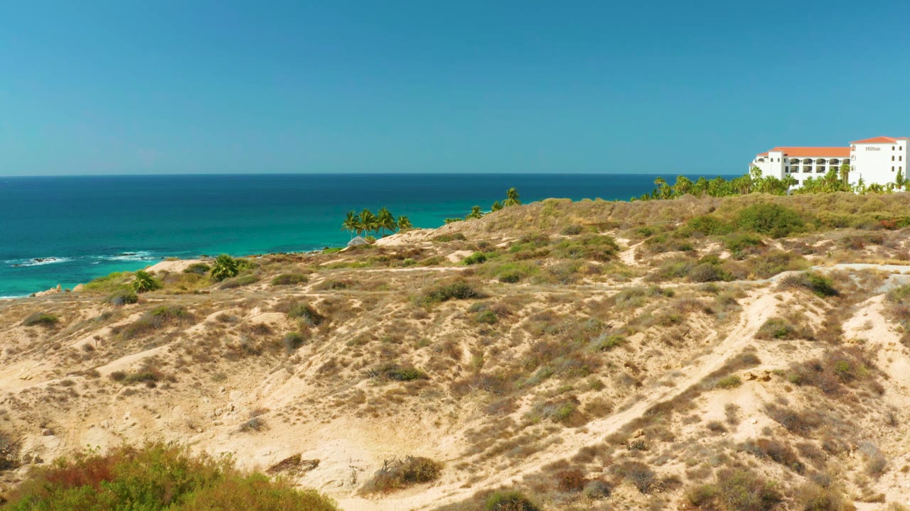 Scrub hillside and turquoise ocean with distant beach palms and building.