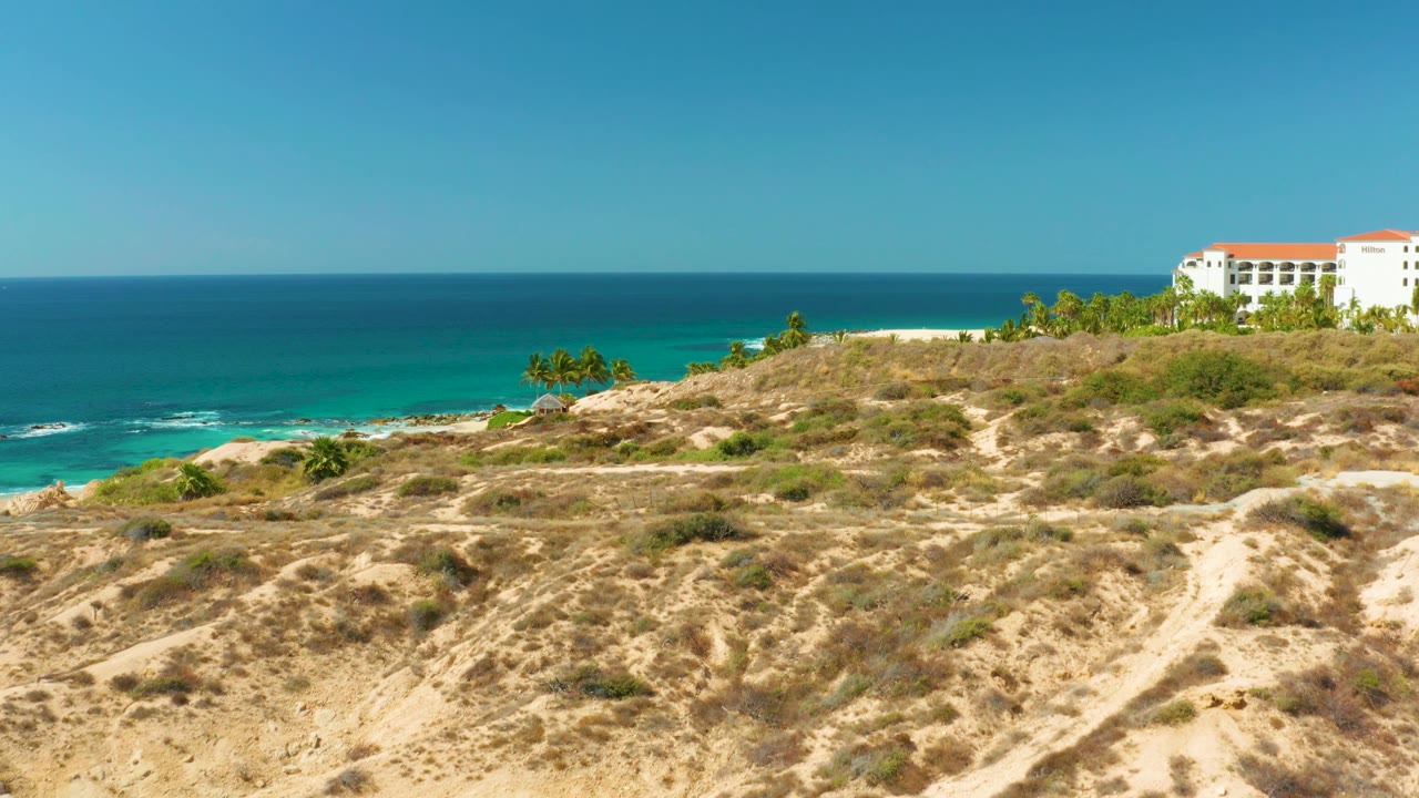 At Hilton Los Cabos BCS Mexico, the coastline begins to open up, showing white sand beyond the dry hillside, palms along the shore, turquoise water, and no visible people.