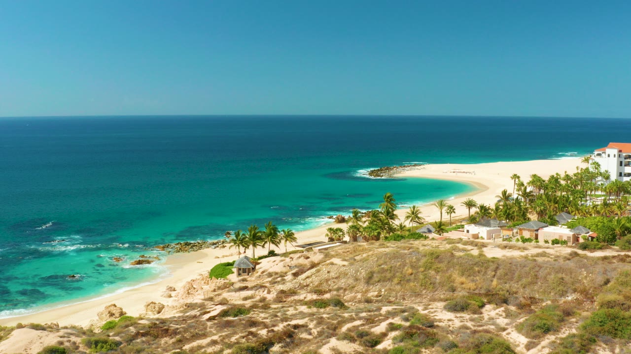 Opening view of Hilton Los Cabos shoreline with turquoise water, pale sand, palm trees, rocky foreground, a taller white building with a red roof, and no visible people.