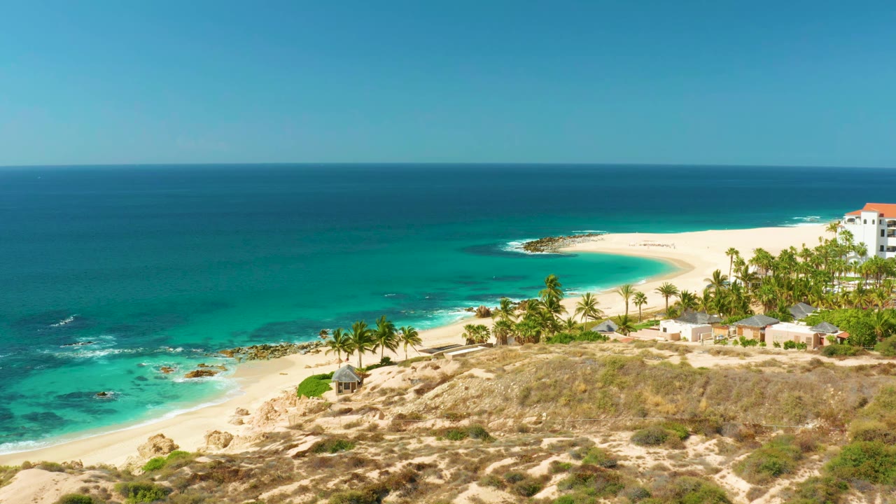 Hilton Los Cabos beach view with the ocean filling the left side, white sand curving around the bay, palms and low buildings on the right, and no visible people.