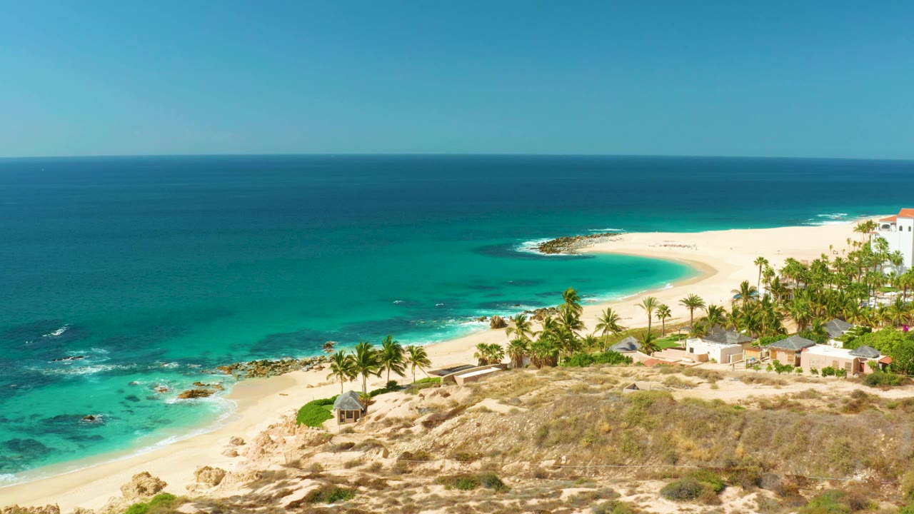 Later view across Hilton Los Cabos beach with more shoreline visible to the right, turquoise water over rocks, palms near the sand, low buildings, and no visible people.