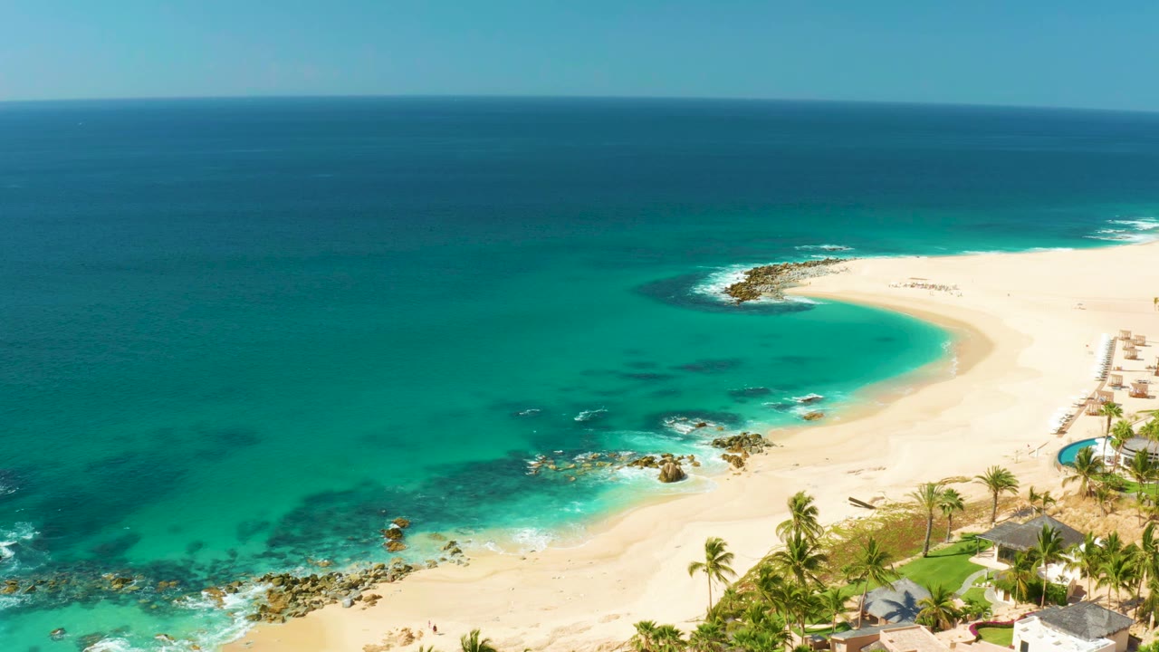 Aerial Hilton Los Cabos coastline view with a broad turquoise bay, curving sand, scattered rocks, palm tops, a narrow resort edge, and no visible people.
