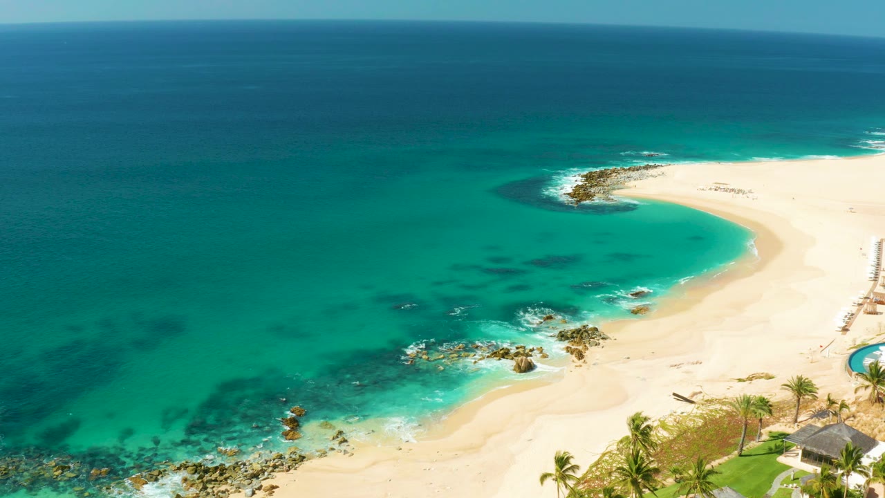 Hilton Los Cabos aerial frame with turquoise ocean taking most of the view, a bright sand arc to the right, rocks in the surf, sparse palm tops, and no visible people.