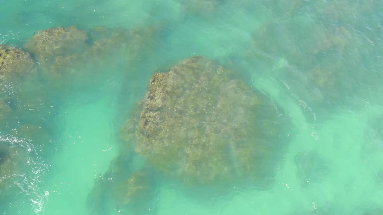 A high overhead frame shows tan submerged rocks under turquoise water at Hilton Los Cabos, Mexico, with faint foam along the left side and no visible people.