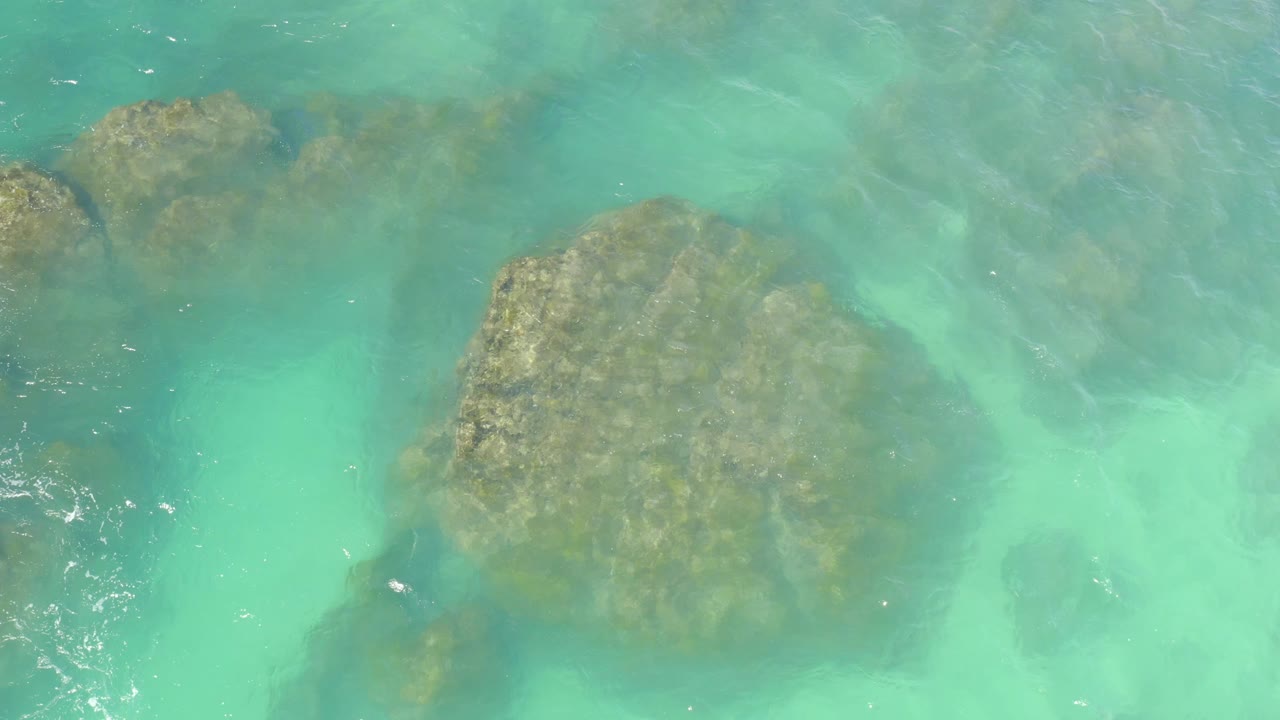 The large underwater rock appears lower in the overhead view as clear turquoise water and soft ripples cover the frame at Hilton Los Cabos, Mexico, with no visible people.
