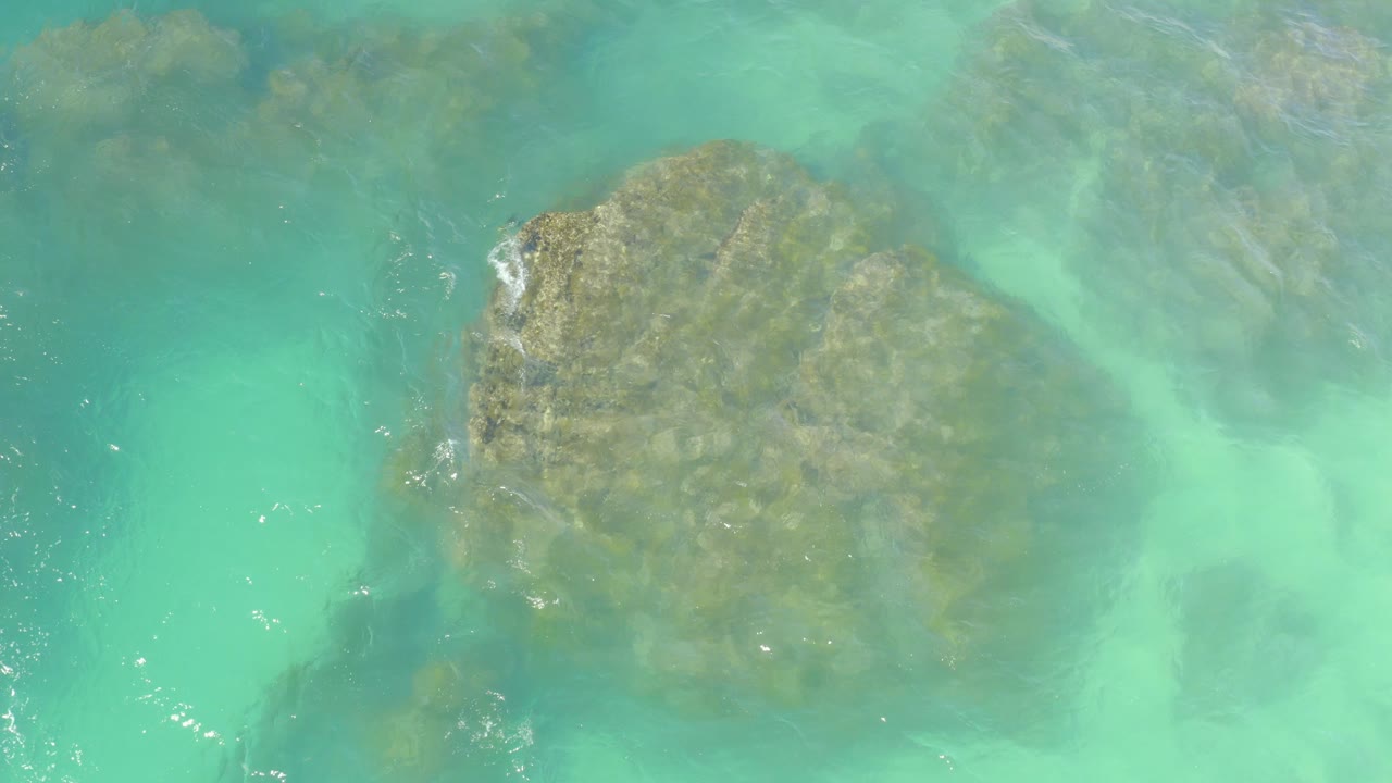 At Hilton Los Cabos in Mexico, clear turquoise water covers a large submerged rock near the center-left of the overhead frame, with no visible people.