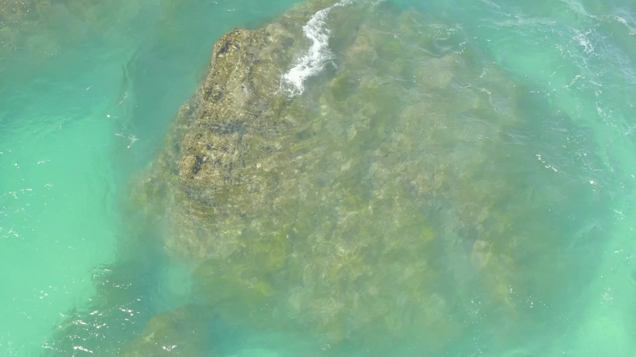 Aerial frame from Hilton Los Cabos showing the submerged rock beneath green-blue water, with white foam touching the upper left edge and no visible people.