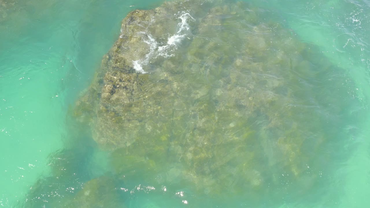 Overhead Hilton Los Cabos view of the same submerged rock through milky turquoise water, with a narrow patch of foam near the upper left and no visible people.