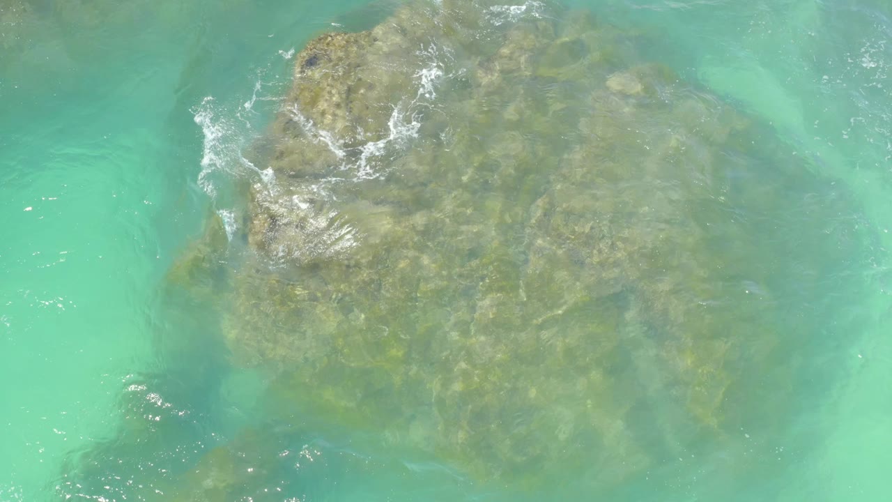 Aerial view at Hilton Los Cabos with turquoise water covering a broad rock, while white foam breaks along the left side and no visible people appear.