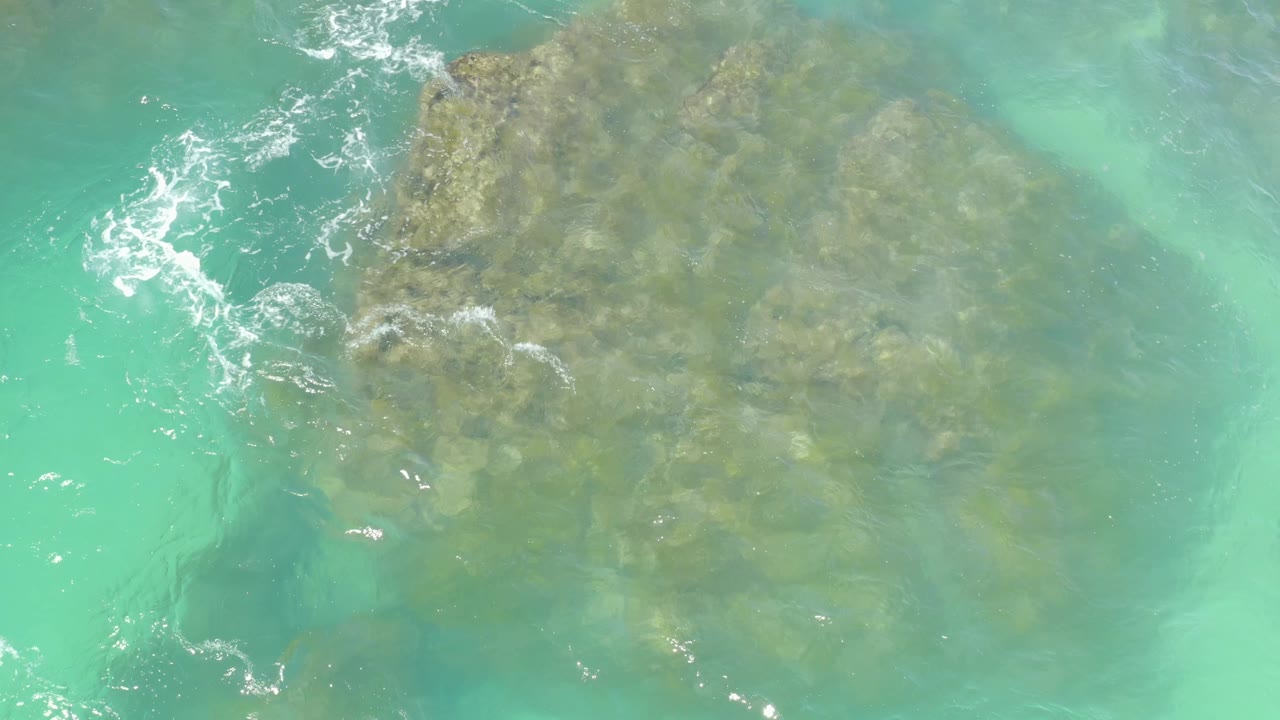 Clear turquoise water at Hilton Los Cabos, Mexico, reveals the rocky seabed, with brighter foam streaks along the left edge and no visible people.