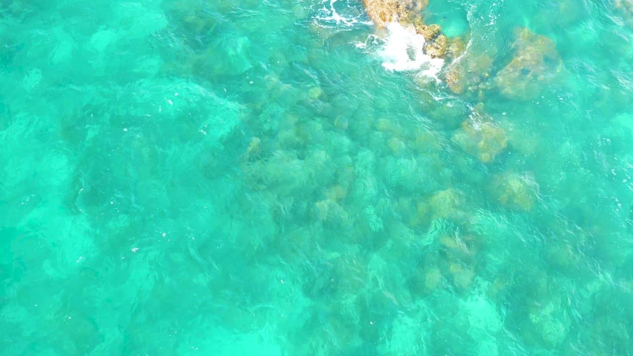 Overhead frame from Hilton Los Cabos, Mexico showing aqua water, darker submerged rocks, and white foam shifting across the upper edge, with no visible people.