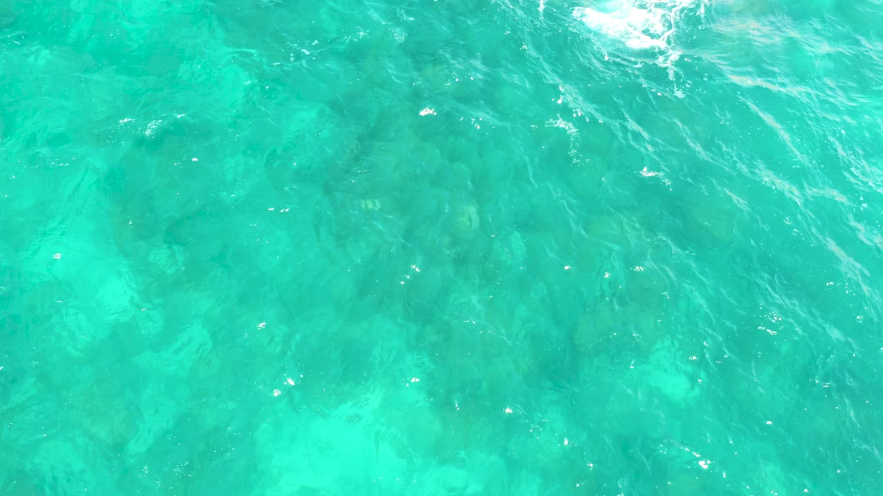 Overhead Hilton Los Cabos, Mexico view of aqua water where submerged rocks sit below rippled surface texture and a small foam patch appears near the upper left, with no visible people.