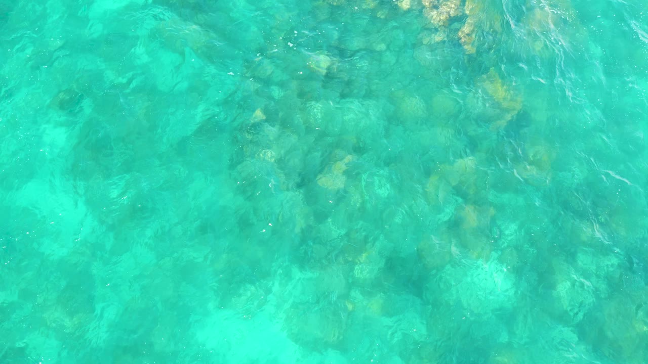 A rocky seabed is visible through rippled turquoise water at Hilton Los Cabos, Mexico, with soft light reflections and no visible people.