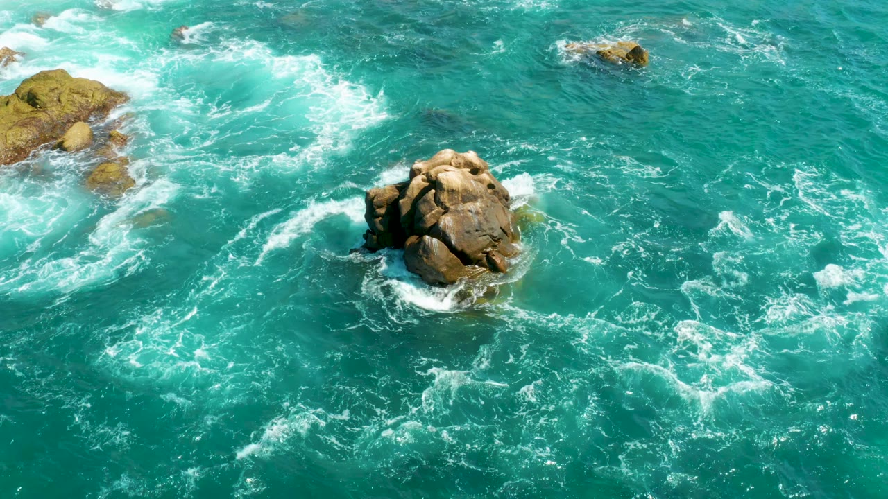 Aerial frame from Hilton Los Cabos, Mexico with the central rock slightly lower in view as foam gathers along its left edge and no visible people.