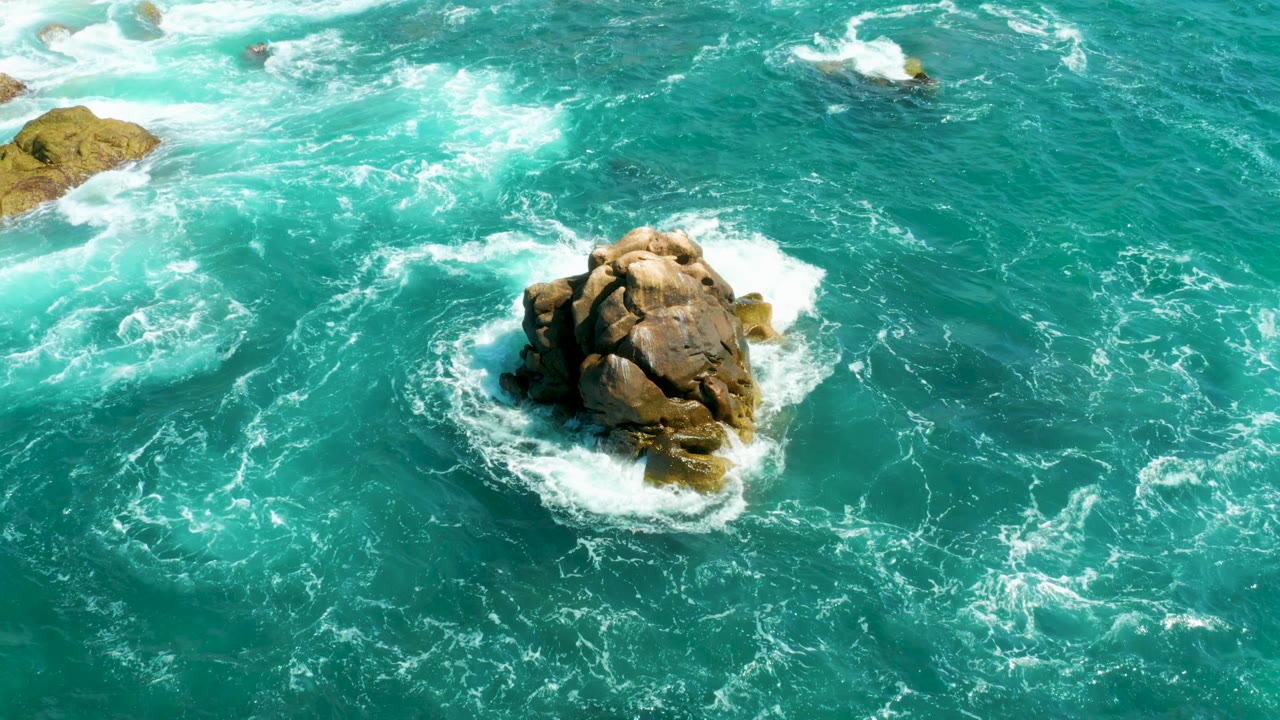 Aerial frame at Hilton Los Cabos, Mexico with white foam wrapping tightly around the central rock and pale submerged rocks visible near the upper left, with no visible people.