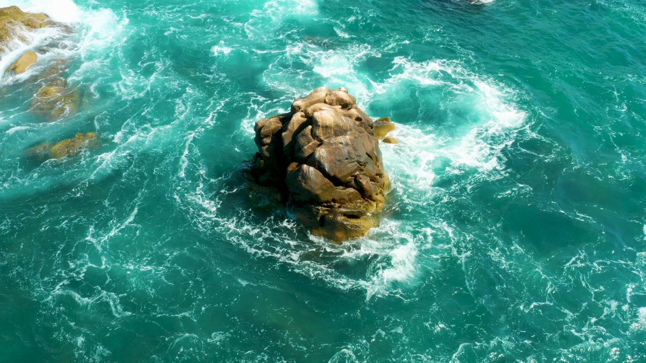 Overhead view at Hilton Los Cabos, Mexico, showing a tan rock outcrop near the center as white foam wraps around it in turquoise water, with no visible people.