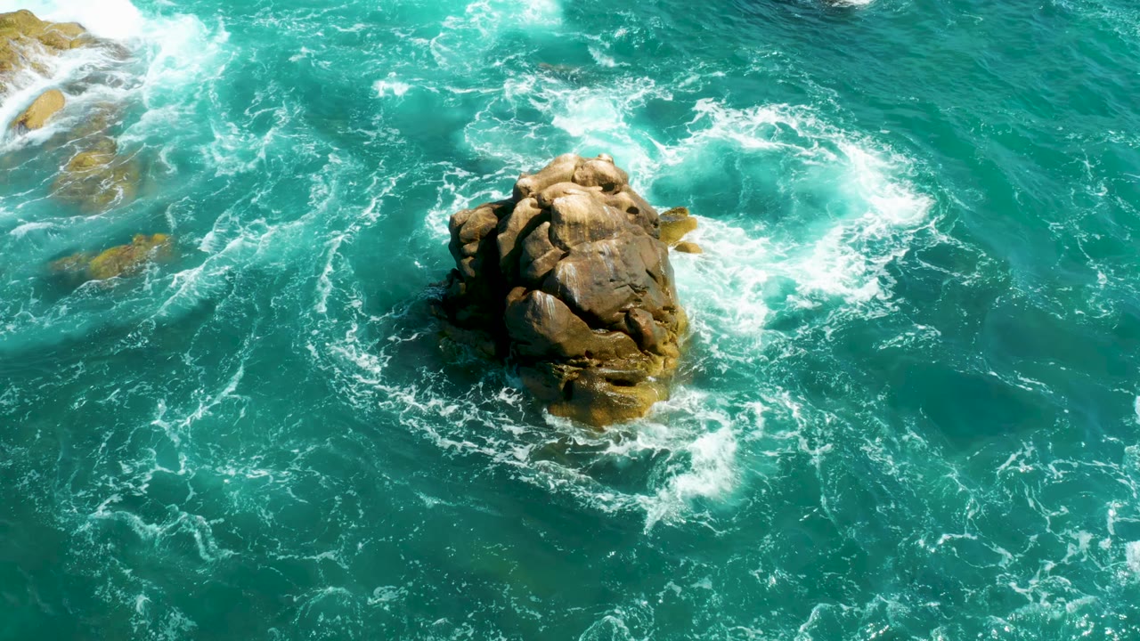 Aerial frame from Hilton Los Cabos, Mexico, with the rock still centered while fresh whitewater spreads across the upper right and lower edge, with no visible people.