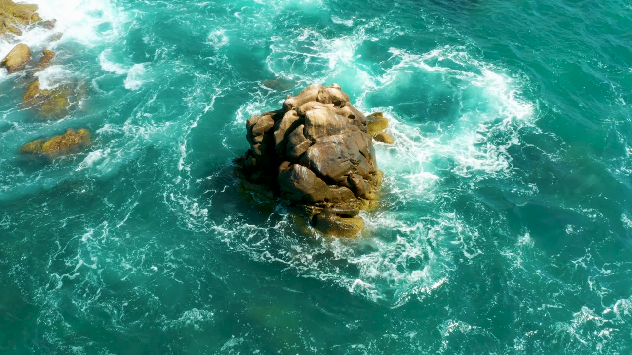 Overhead Hilton Los Cabos ocean view with the rock slightly right of center, heavy foam above it, and scattered submerged stones along the left edge, with no visible people.