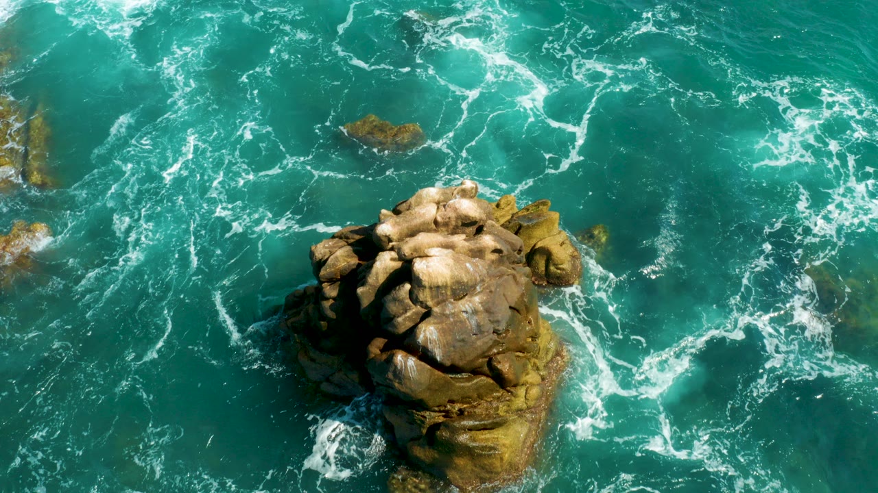 Overhead view of a tan sea rock surrounded by turquoise water and white foam.